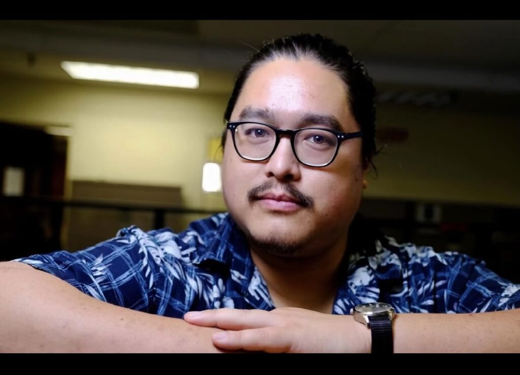 Man with black glasses, blue shirt and hair pulled back.